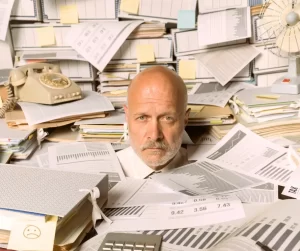Image of a man with an annoyed look on his face drowning in in office room full of paper to represent the overload and confusion involved in understanding health insurance plans.